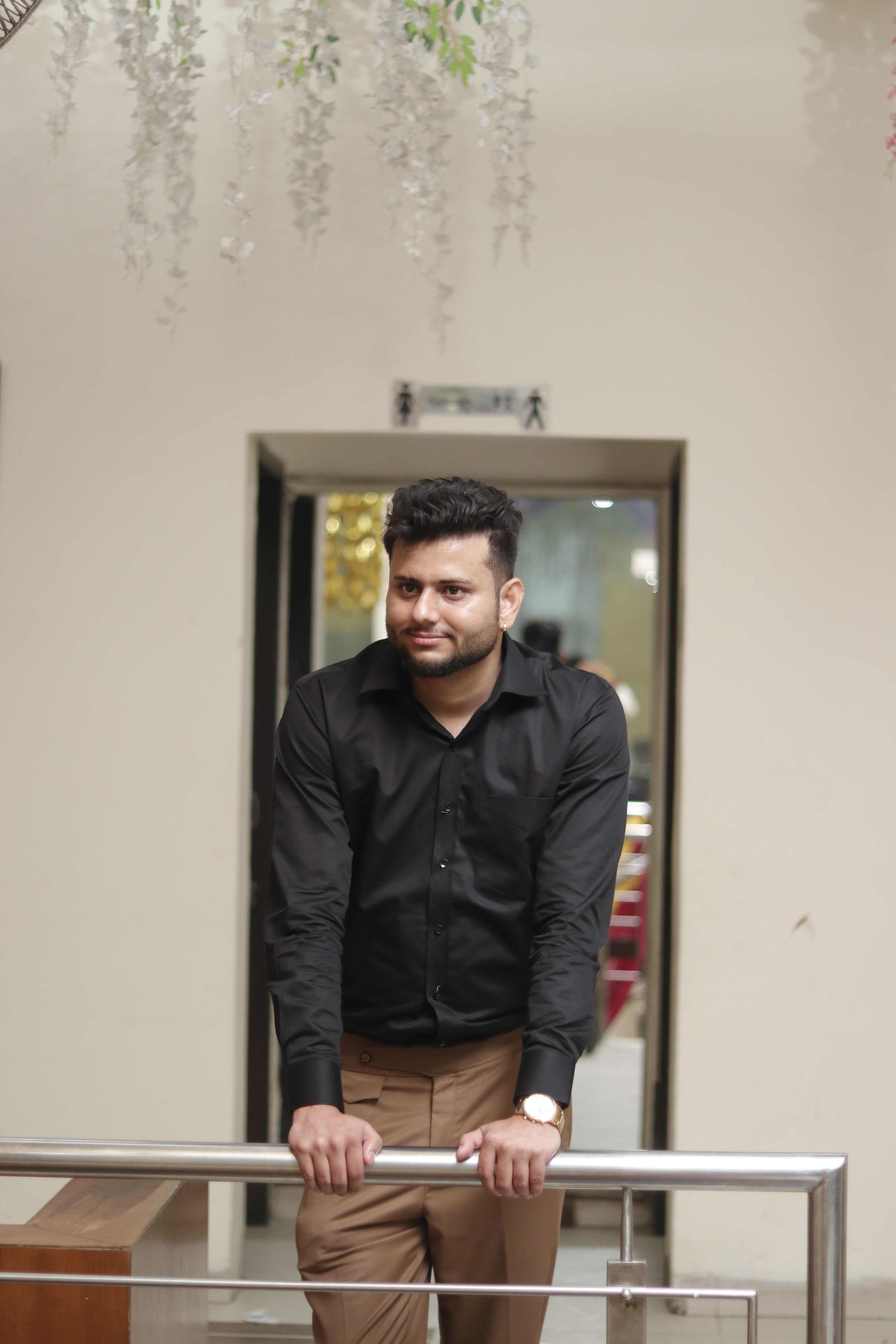 Shubham Verma in a black shirt and tan trousers leaning on a metal railing